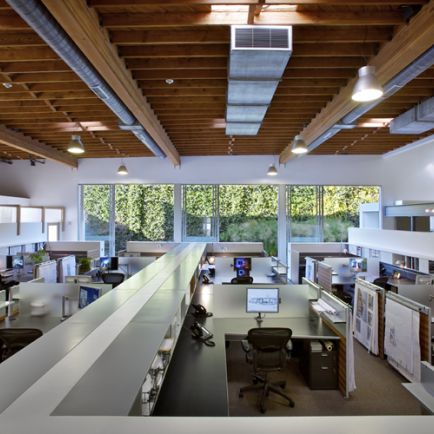 An office space featuring numerous desks arranged throughout, complemented by large windows allowing ample natural light.