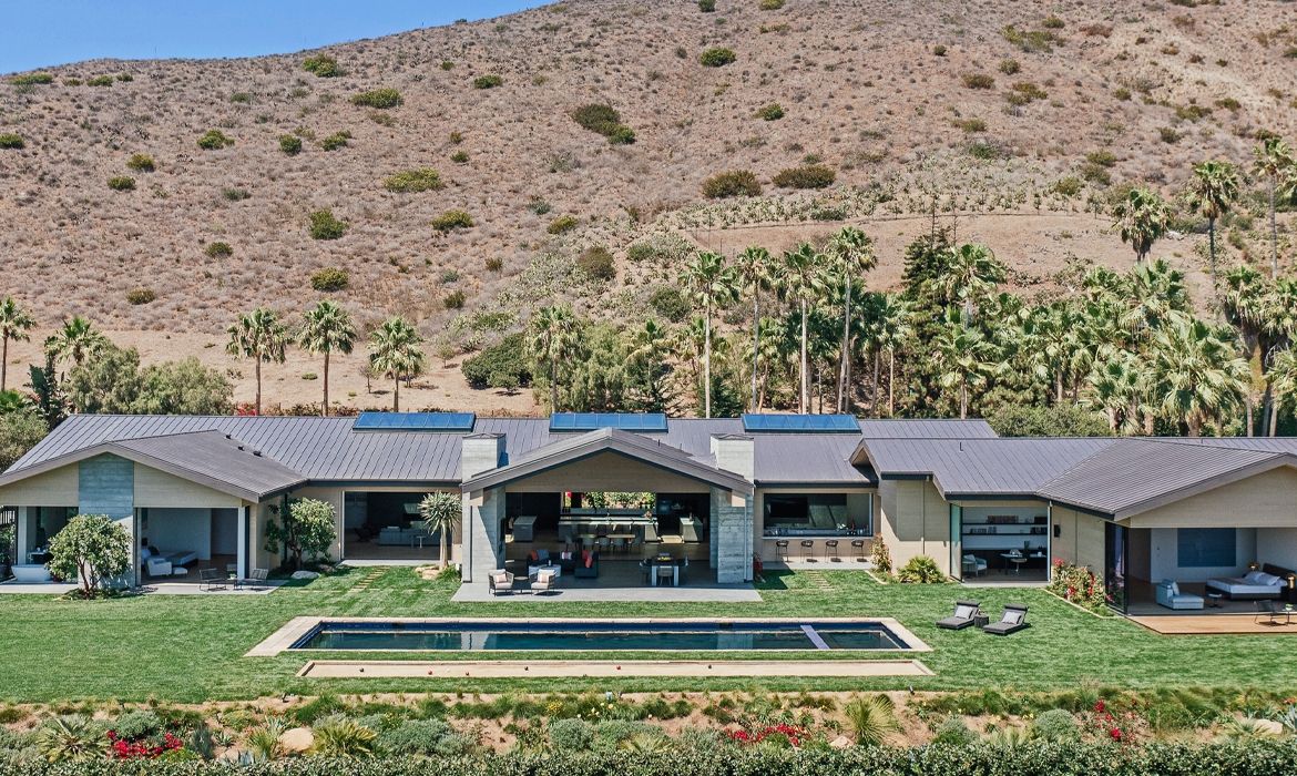 Modern hillside villa with solar panels, a central pool, and surrounding palm trees. Open-air design with expansive glass walls and green lawn. Desert landscape in the background.