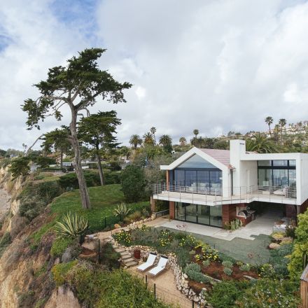 Modern two-story house perched on a coastal cliff, surrounded by lush greenery and a large tree, with a view of the ocean.